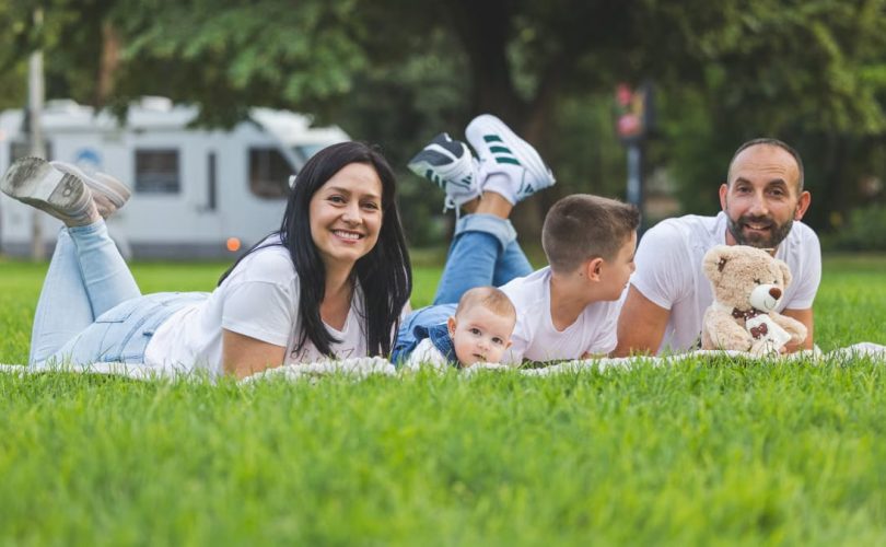 Happy family lying on grass with RV in the background, enjoying a sunny outdoor picnic.