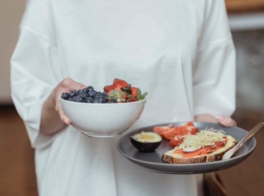 Person holding a bowl of fresh berries and a plate with avocado toast, displaying a healthy breakfast spread.